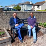Two people sitting on a bench in a community green space.